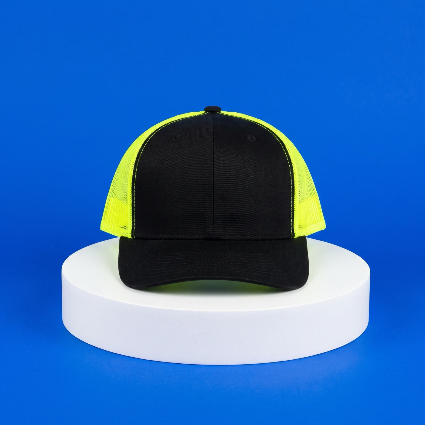 Black trucker hat with neon yellow mesh on a white pedestal against a bright blue background