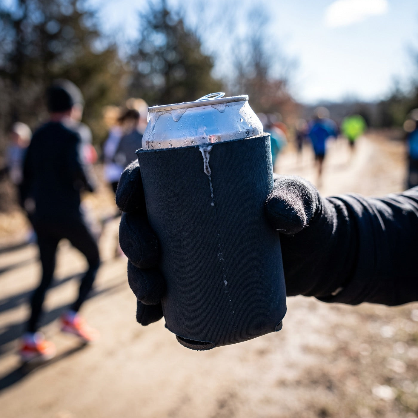 Hand gripping a frosty can in a black neoprene koozie with a race blurred in the background